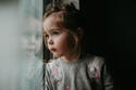 Little girl wearing grey sweater leaning on window in shadow
